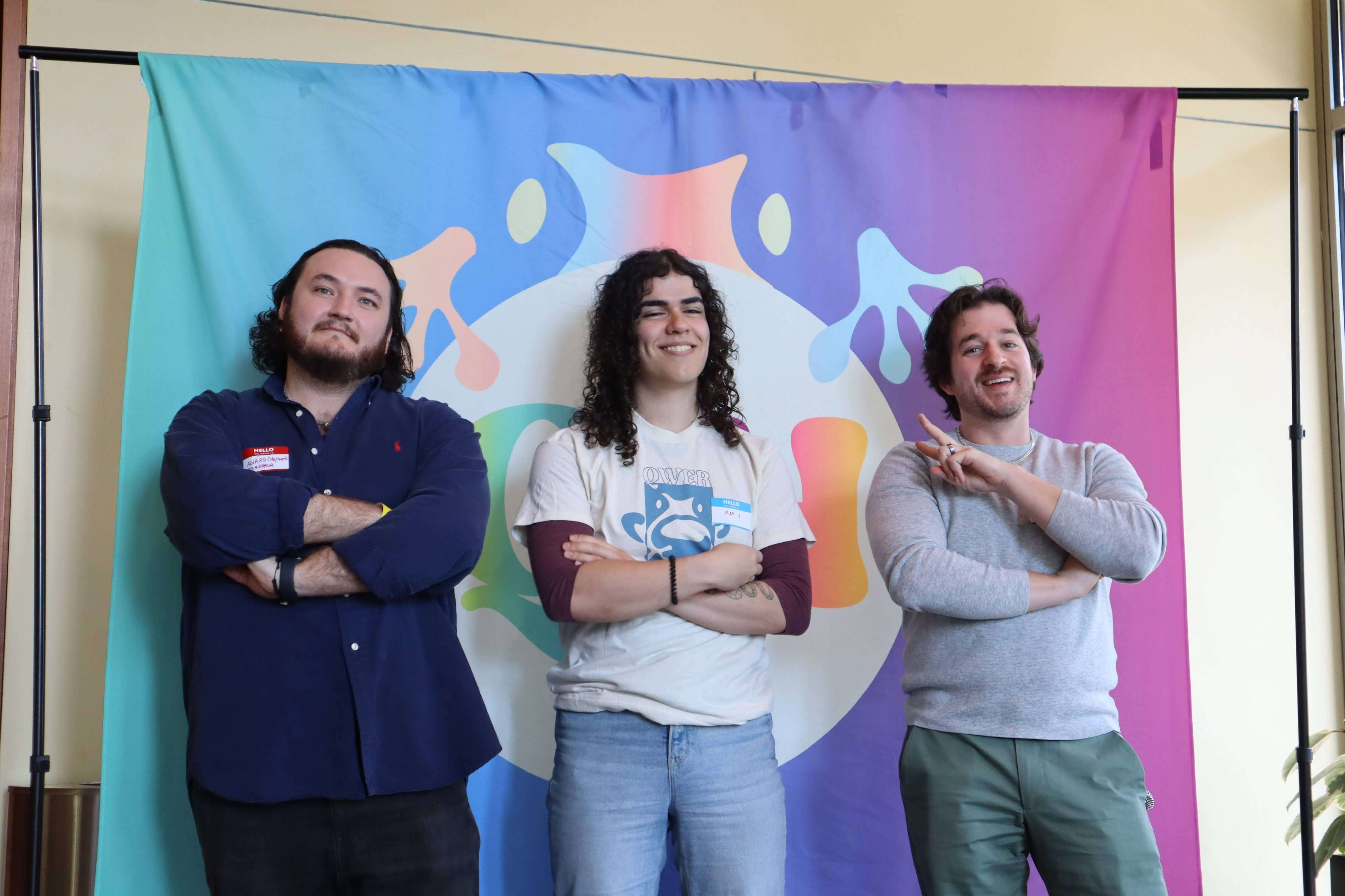 Three people posing with crossed arms in front of a colorful banner with abstract designs. From left to right: a person with a beard and long hair, wearing a navy shirt; a person with curly shoulder-length hair in a white T-shirt with a blue frog graphic; and a person with short wavy hair in a grey sweater. They all appear cheerful.